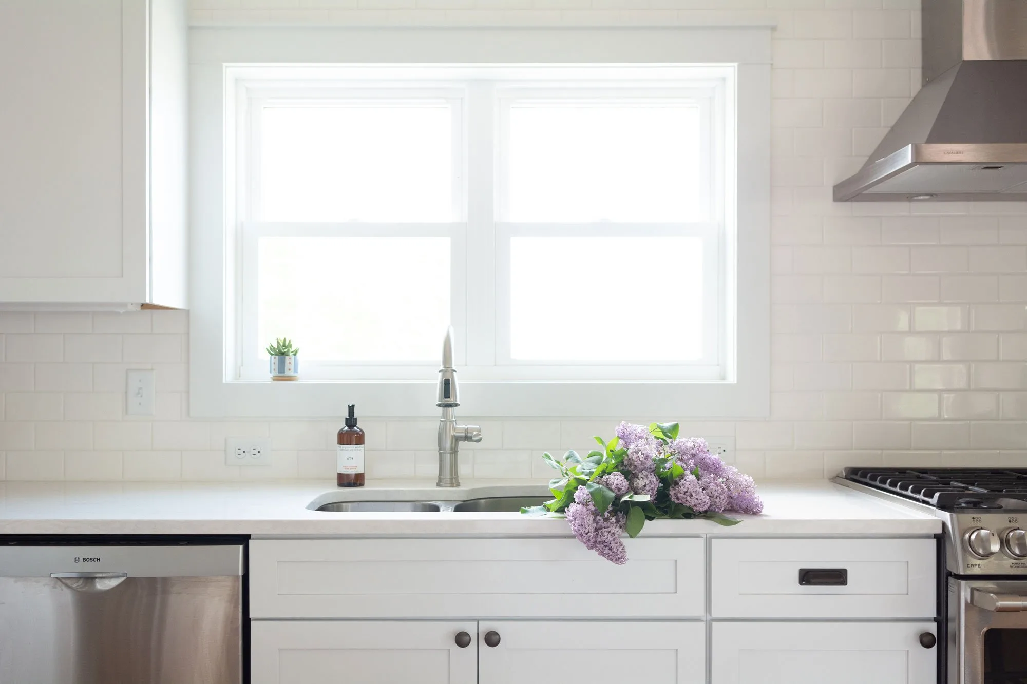 White kitchen sink below a window, with lilacs on the counter